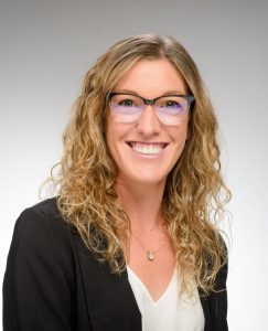Brooke has shoulder-length light brown, curly hair, and she is smiling and wearing glasses, a white shirt, and black blazer