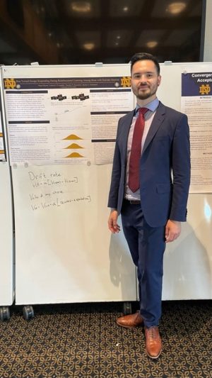 Miguel has short, dark brown hair and is standing in front of his research poster, smiling and wearing a blue suit with a red tie and light brown shoes