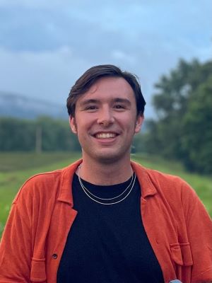 Connor has short brown hair and he is smiling outside while wearing a black shirt and orange jacket