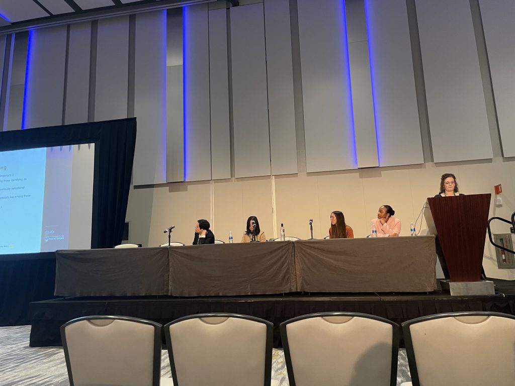 Members of the ASSIST Lab sit at the front of a large group at a long table looking at a presentation on a screen to their right