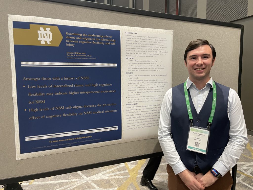 Connor stands smiling in a white shirt and blue sweater vest in front of his research poster