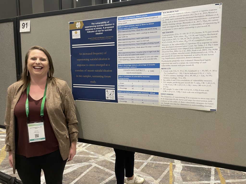 Anne stands in front of her research poster wearing a dark red shirt, tan blazer, and black pants, smiling