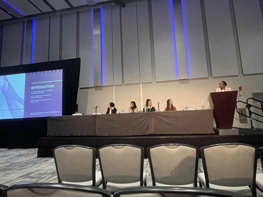 Members of the ASSIST Lab sit at the front of a large group at a long table looking at a presentation on a screen to their right in a different position