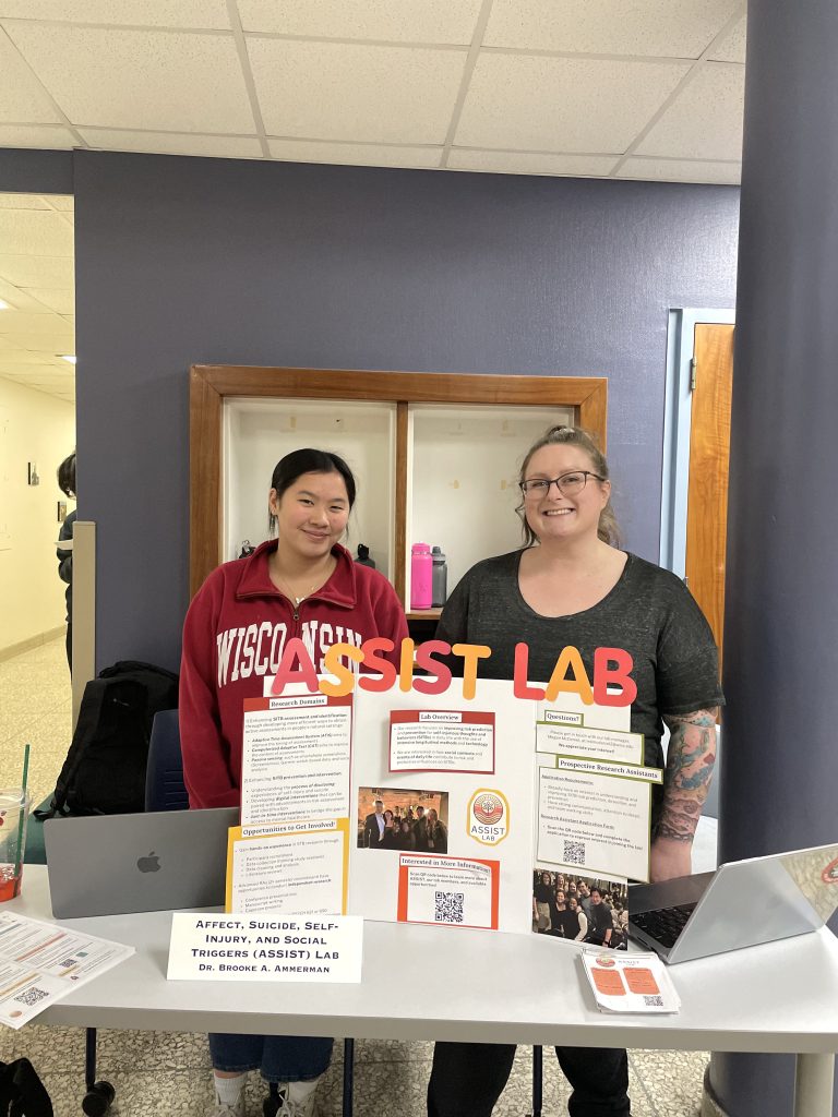 Megan stands next to Jenna behind the ASSIST Lab poster.