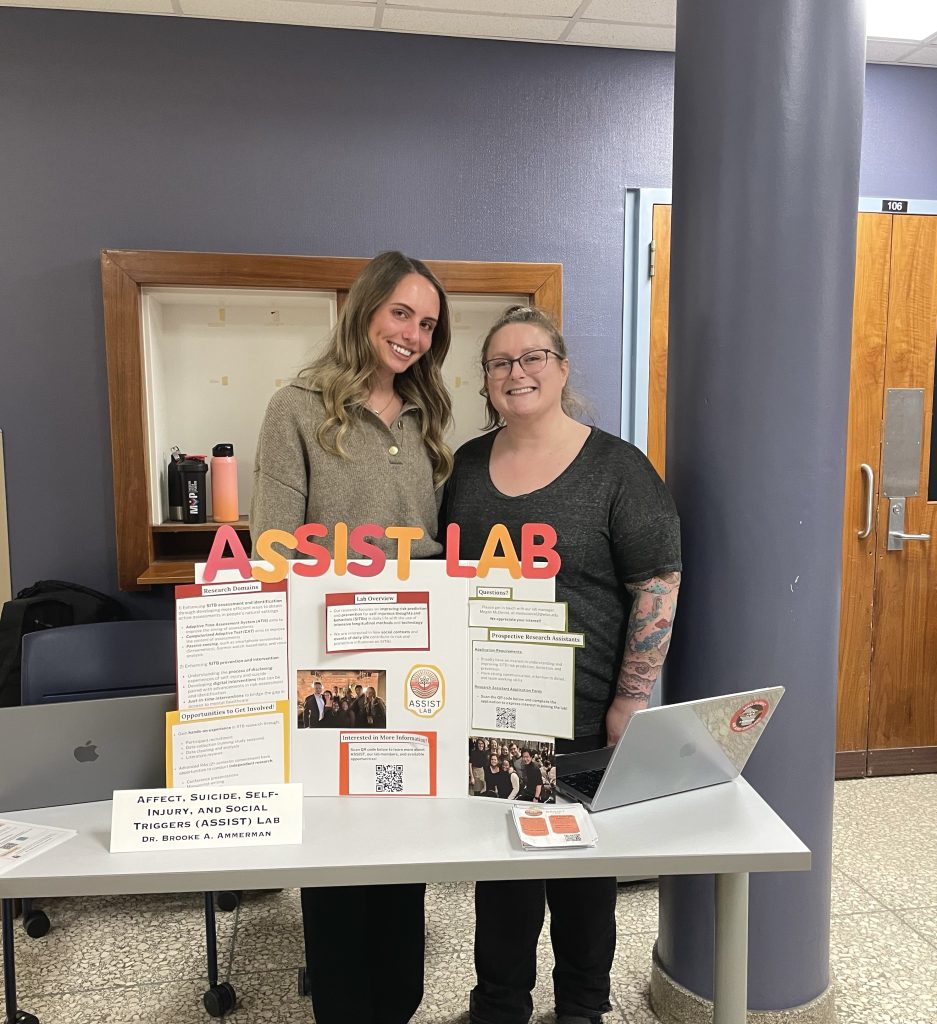 Megan is wearing a dark gray shirt standing next to Veronika wearing a tan sweater. They are standing behind an ASSIST Lab poster on a table.