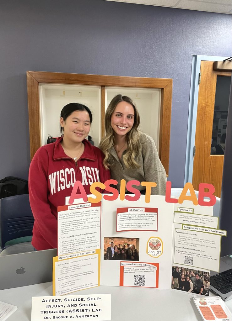 Veronika, smiling and wearing a tan sweater, stands next to Jenna, wearing a red Wisconsin sweater. They are standing behind the same ASSIST Lab poster.