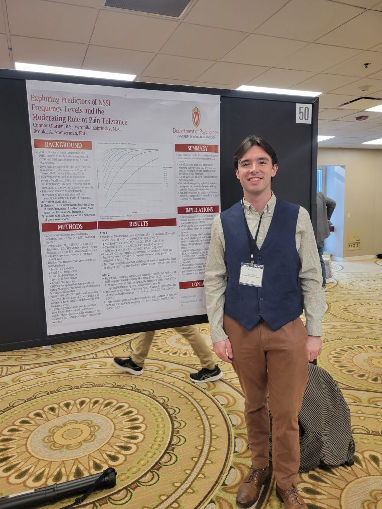 Connor stands smiling in front of his research poster wearing a green shirt, dark blue sweater vest, brown pants, and brown dress shoes