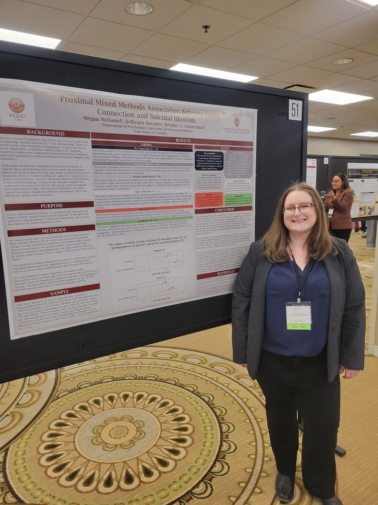 Megan stands in front of her research poster, smiling and wearing a dark blue shirt, gray blazer, and black pants, standing on a patterned rug