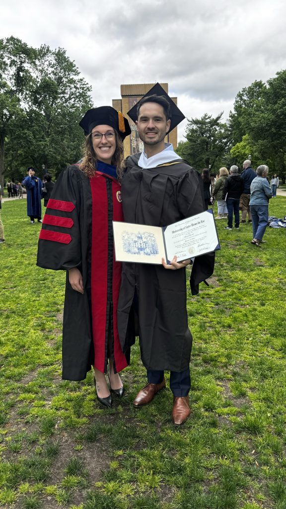 Image of Brooke and Michael smiling outside wearing graduation regalia with Michael holding open his diploma