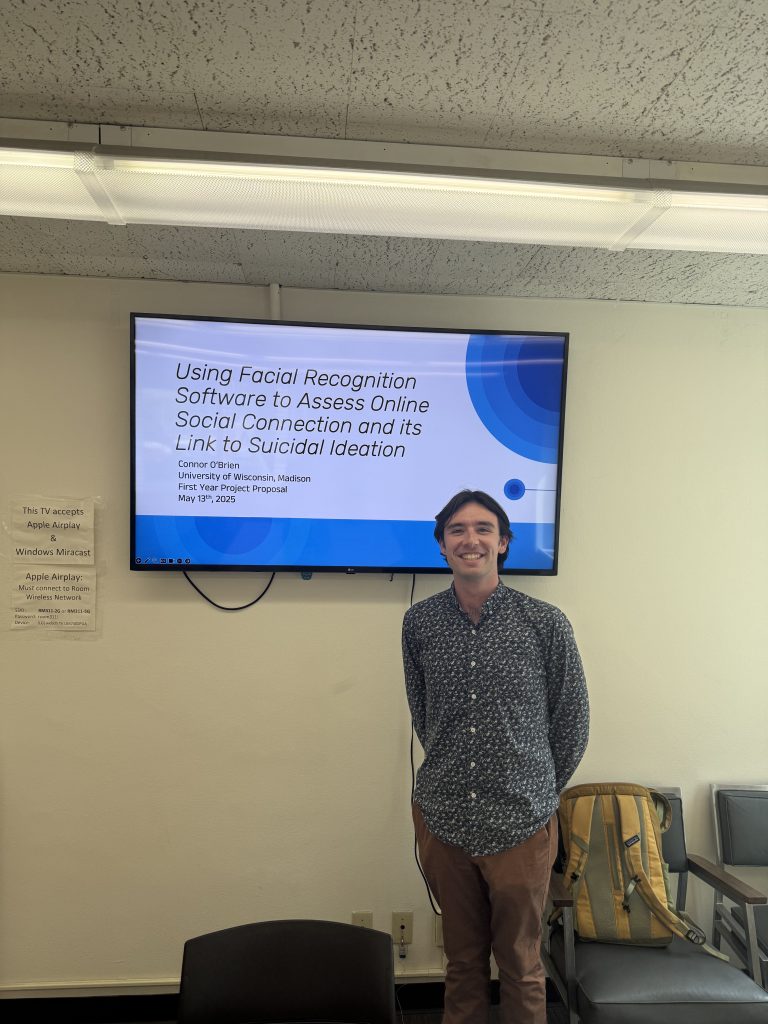 Connor stands in front of a TV smiling, wearing a blue shirt and tan pants and displaying his first-year project presentation