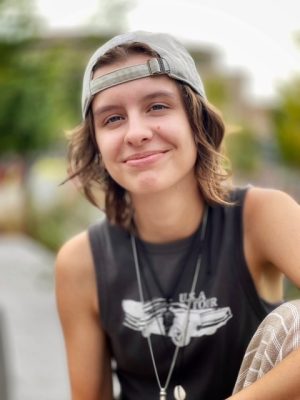 Image of Maya outside wearing a baseball hat backwards in a dark gray tank top, smiling