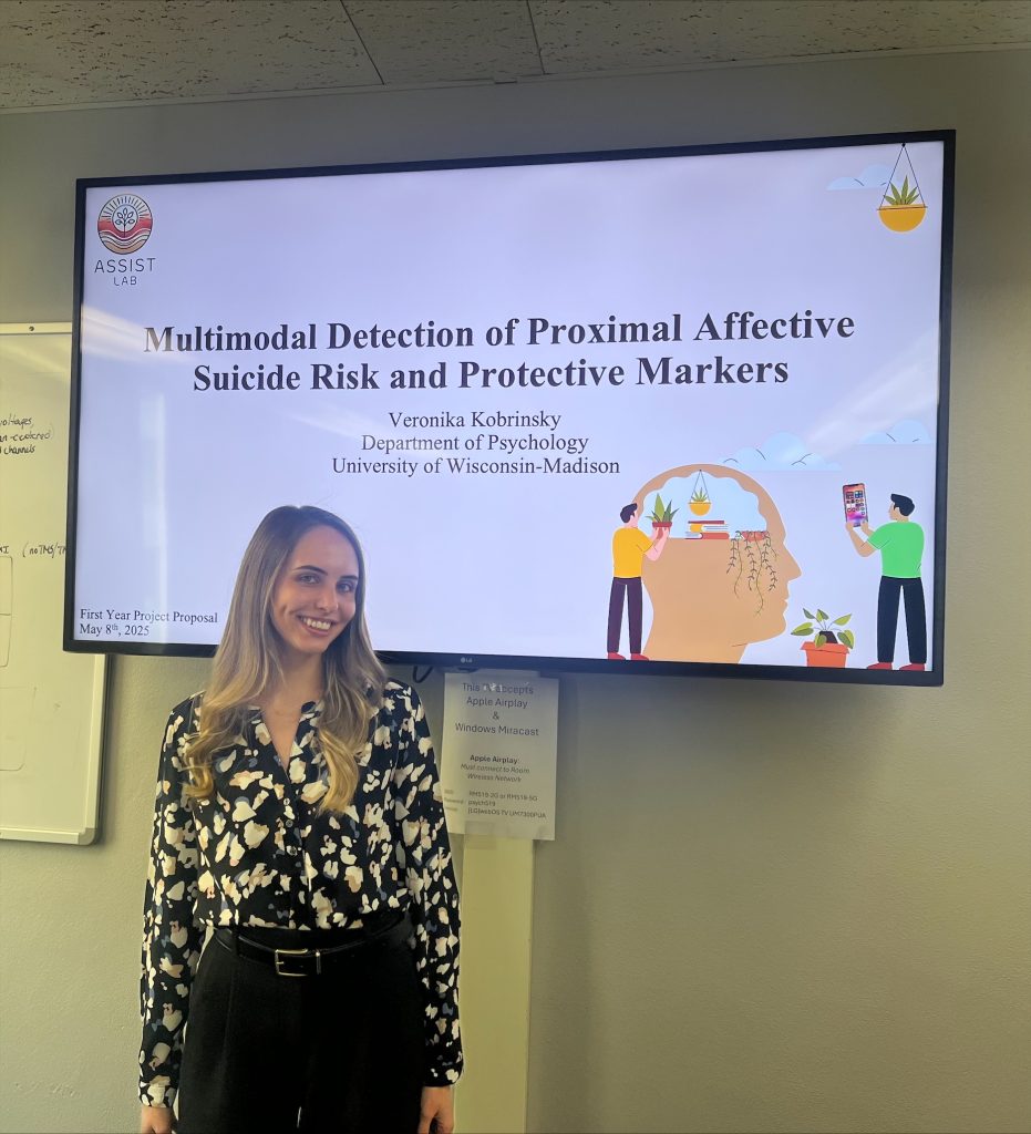 Veronika stands in front of a TV smiling, wearing a white and black shirt and black pants and displaying her first-year project presentation