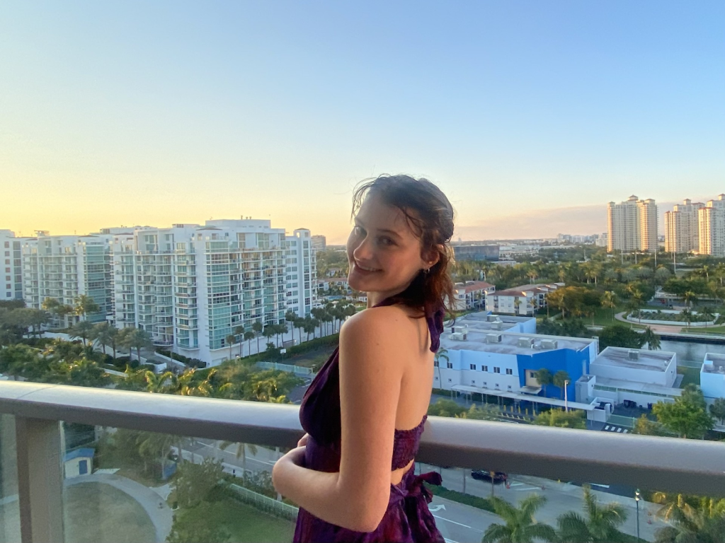 Lily has shoulder-length brown hair and is on a balcony with a city scape behind her, smiling and wearing a purple dress