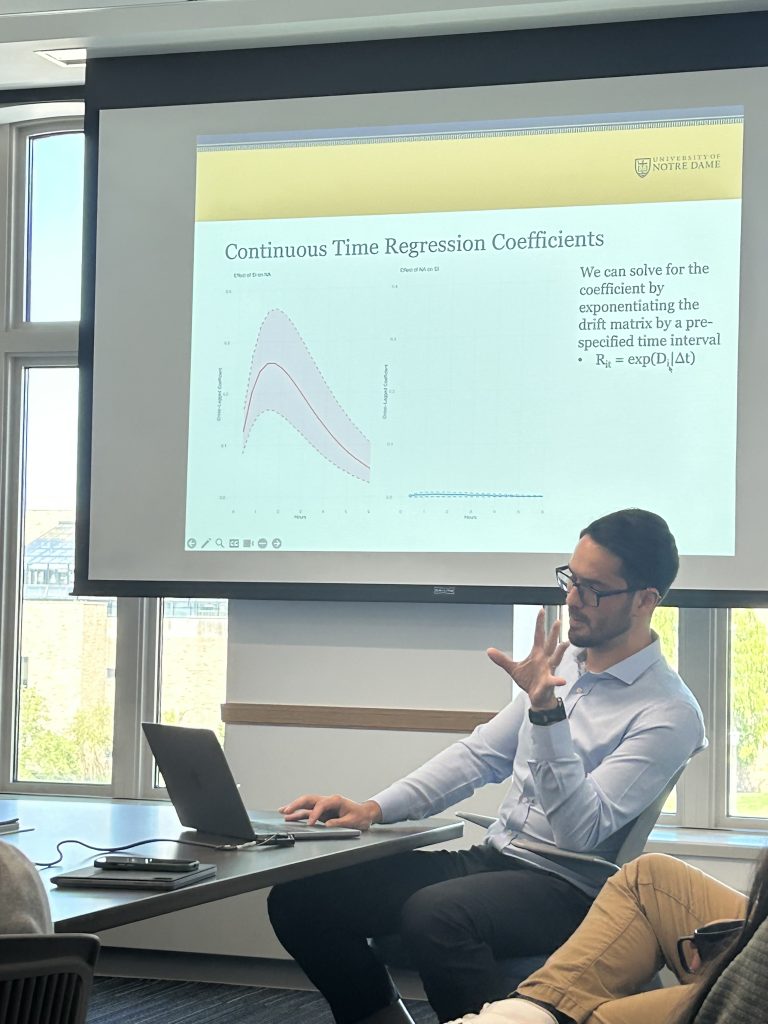 Second picture of Michael sitting in a classroom wearing a light blue shirt and black pants and giving a presentation with his laptop open in front of him
