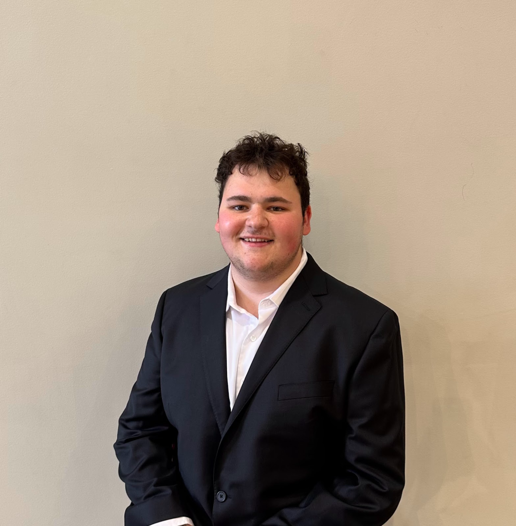 Jake has short, dark brown hair and is smiling wearing a black suit