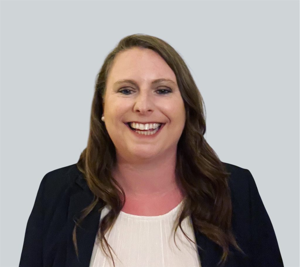 Anne has medium-length dark brown hair and is smiling while wearing a white shirt and black blazer