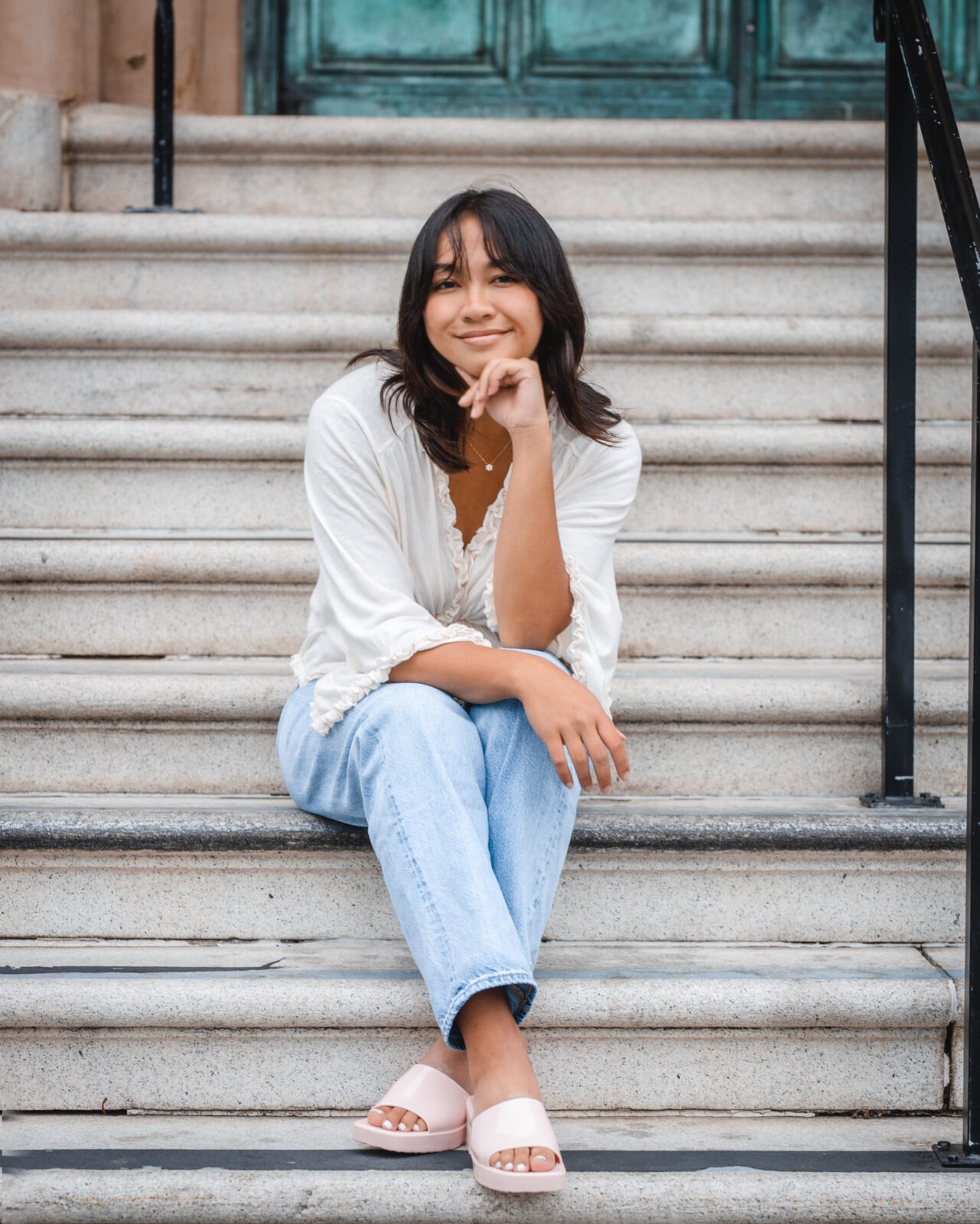 Julia is sitting on steps and is wearing a white shirt with jeans, smiling.