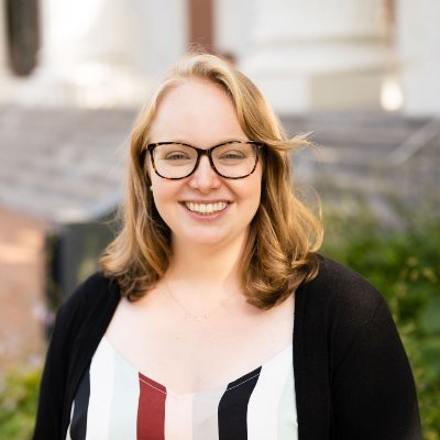 Emily has glasses and is smiling outside.
