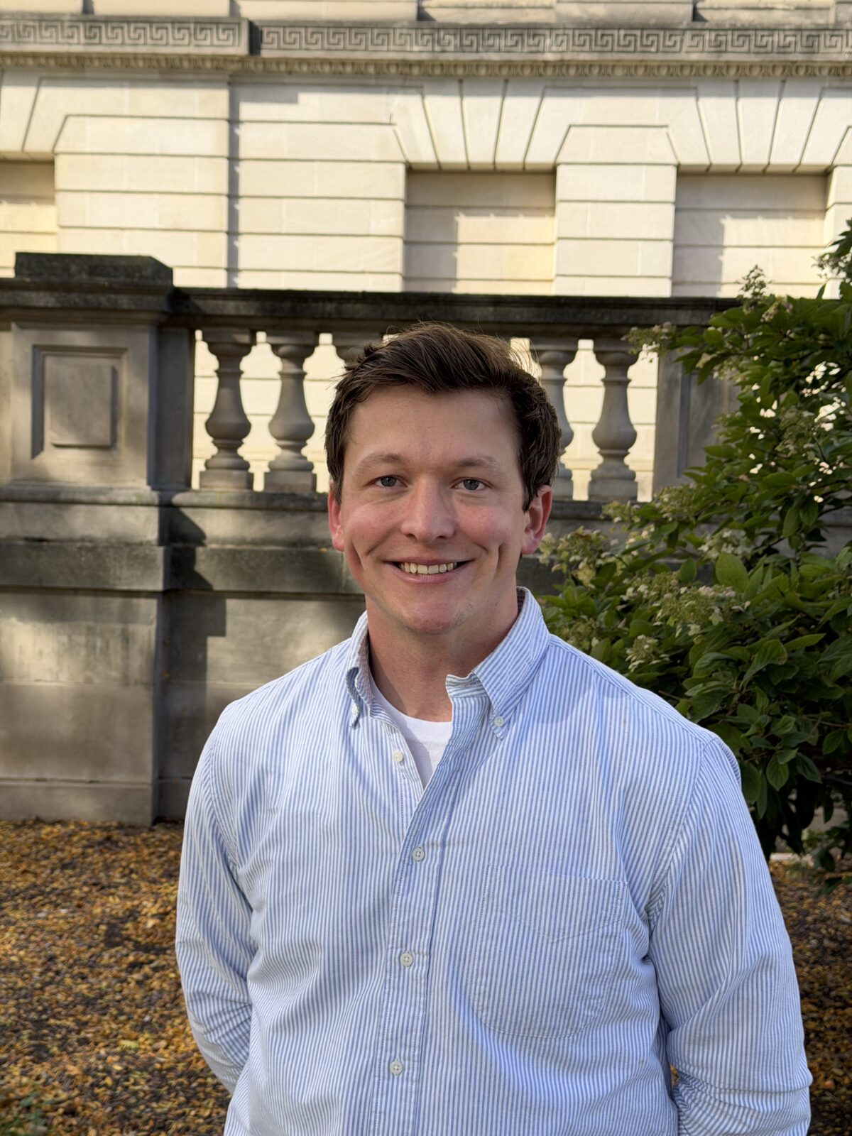 Nick is smiling and is standing outside in a blue shirt.